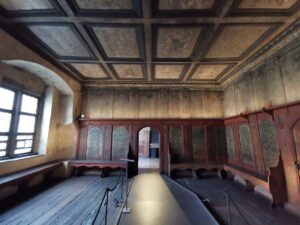 Umlaufenden, dunkel getäfelten Holzwände mit integrierten Sitzbänken sowie die bemalte Kassettendecke aus Holz